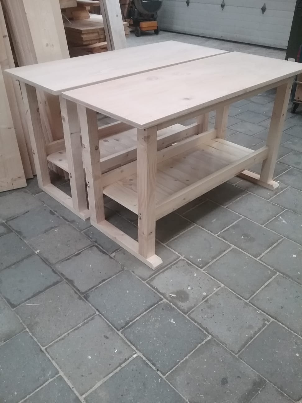 Twee nieuwe tafels in blank hout met een legplank onderin, op de bestrating voor de werkplaats
